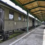Po lewej schodki do zabytkowego wagonu pociągu stojącego przy zadaszonym peronie z namalowaną białą linią. Na peronie wzdłuż stoją ławki. Na jednej z nich siedzą ludzie.